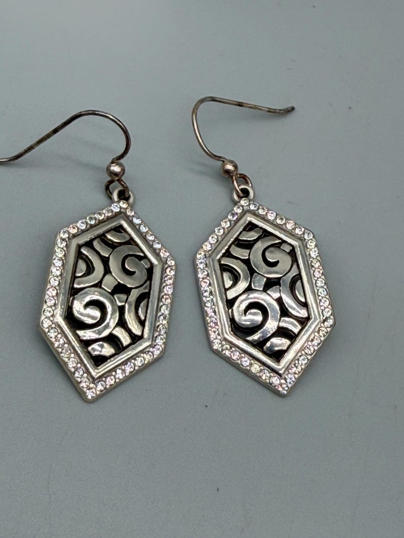 Puede incluir: Un par de pendientes colgantes plateados. Cada pendiente presenta un dise&ntilde;o geom&eacute;trico calado con un patr&oacute;n de remolino, enmarcado por un borde de peque&ntilde;os cristales transparentes. Cierre de gancho.