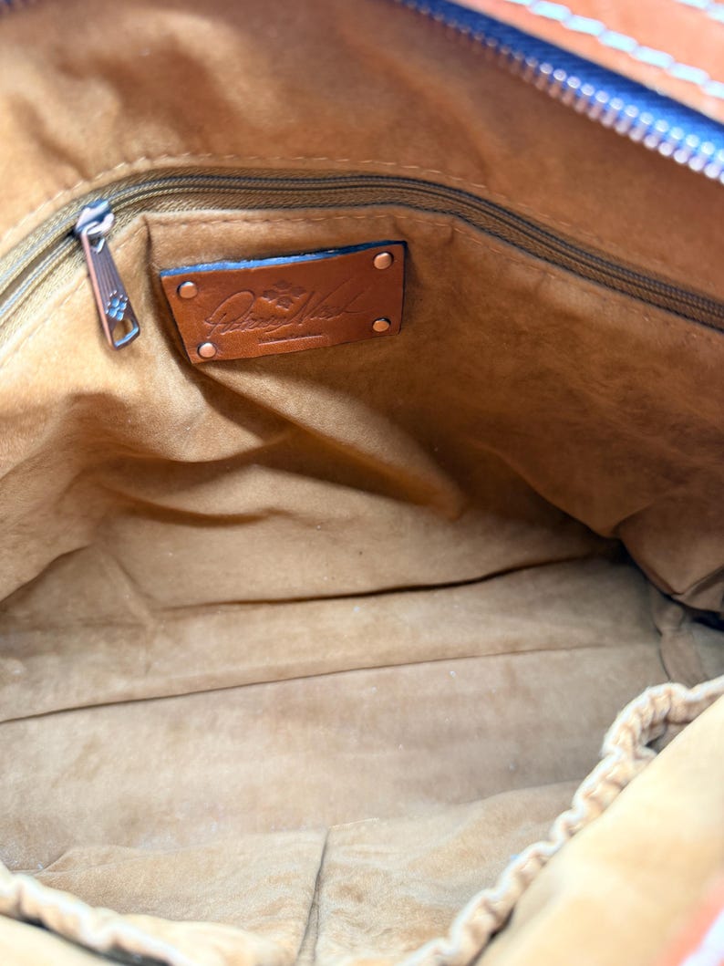 May include: Interior view of a brown leather handbag. The inside is lined with a soft, light brown material. A zipper runs along the top, and a leather tag with a logo is visible. The bag is open, showing the spacious interior.