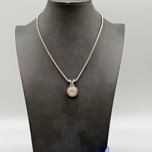 May include: A silver-toned rope chain necklace featuring a circular pendant. The pendant has a central clear stone surrounded by a gold-toned inner ring and a textured silver-toned outer border with a twisted rope design and a decorative bail.
