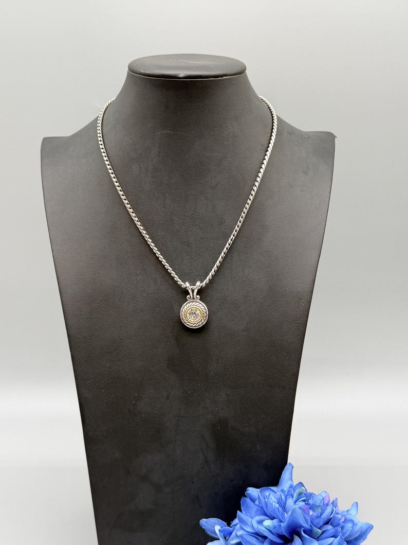 May include: A silver-toned rope chain necklace featuring a circular pendant. The pendant has a central clear stone surrounded by a gold-toned inner ring and a textured silver-toned outer border with a twisted rope design and a decorative bail.