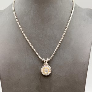 May include: A silver rope-style chain necklace featuring a circular pendant. The pendant has a central clear stone surrounded by a gold-toned inner ring and a silver-toned outer border with a twisted rope texture and decorative metal accents.