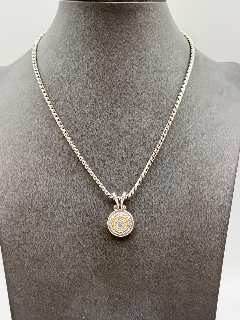 May include: A silver rope-style chain necklace featuring a circular pendant. The pendant has a central clear stone surrounded by a gold-toned inner ring and a silver-toned outer border with a twisted rope texture and decorative metal accents.