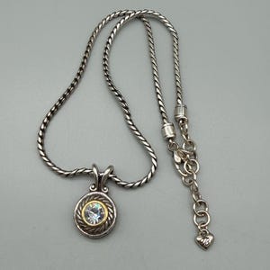May include: A silver-toned necklace featuring a twisted rope chain and a circular pendant. The pendant has a central clear faceted stone surrounded by a gold-toned rim and a silver-toned rope border. An adjustable chain end features a small heart charm.