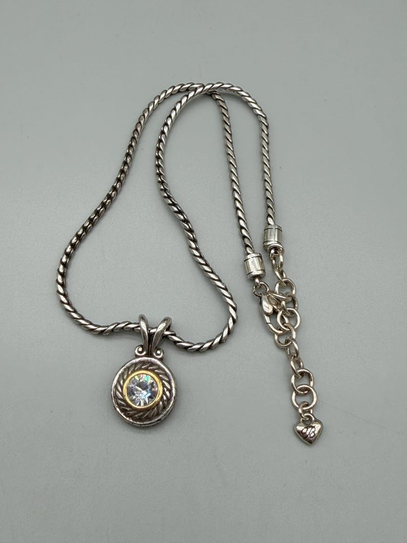 May include: A silver-toned necklace featuring a twisted rope chain and a circular pendant. The pendant has a central clear faceted stone surrounded by a gold-toned rim and a silver-toned rope border. An adjustable chain end features a small heart charm.