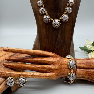 May include: A gold-toned necklace, bracelet, and earrings set. Each piece features silver-toned, circular pendants with an intricate design. The jewelry is displayed on a wooden bust and hand, showcasing the set's details and style.