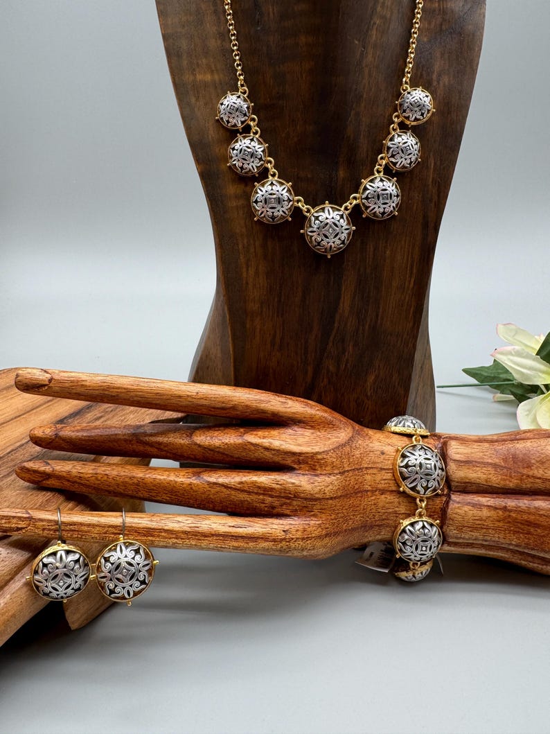 May include: A gold-toned necklace, bracelet, and earrings set. Each piece features silver-toned, circular pendants with an intricate design. The jewelry is displayed on a wooden bust and hand, showcasing the set's details and style.