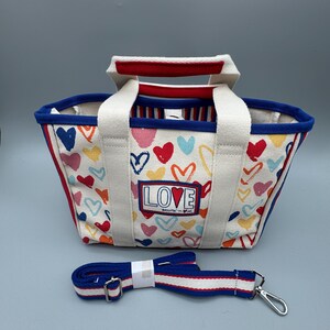 Puede incluir: Un bolso tote de lona con un estampado de corazones coloridos y la palabra "LOVE" en la parte delantera. Tiene asas blancas y una correa de hombro azul desmontable con rayas rojas y blancas. El ribete del bolso es rojo y azul.