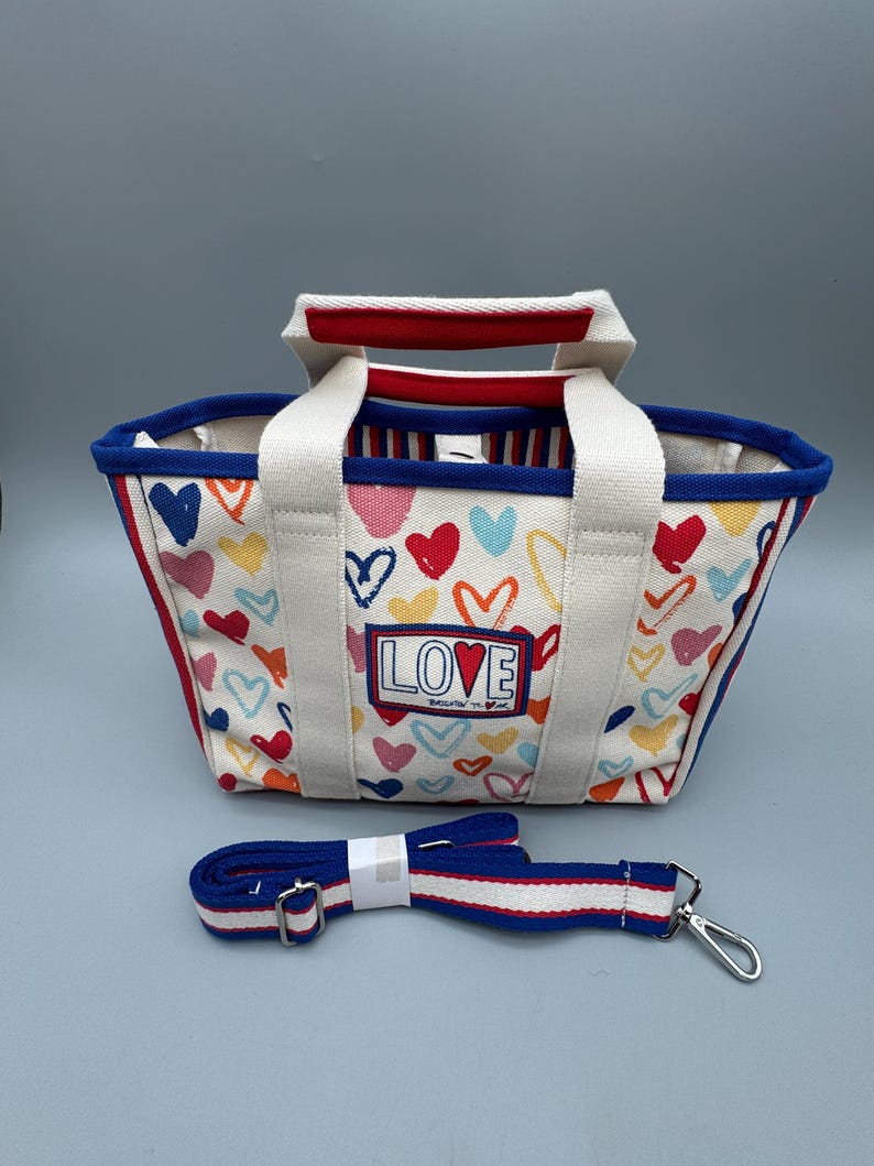 Puede incluir: Un bolso tote de lona con un estampado de corazones coloridos y la palabra "LOVE" en la parte delantera. Tiene asas blancas y una correa de hombro azul desmontable con rayas rojas y blancas. El ribete del bolso es rojo y azul.