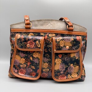 May include: A floral print tote bag with two front pockets. The bag features a dark background with a colorful floral pattern, brown leather trim, and two top handles. The bag has a gray suede-like top.