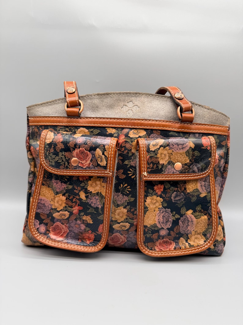 May include: A floral print tote bag with two front pockets. The bag features a dark background with a colorful floral pattern, brown leather trim, and two top handles. The bag has a gray suede-like top.