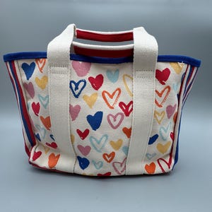 Puede incluir: Un peque&ntilde;o bolso de mano de lona con un estampado de corazones coloridos sobre un fondo blanco. Tiene asas blancas, un ribete azul y rayas rojas y azules en los laterales. Los corazones son de varios colores, como rojo, rosa, azul y amarillo.