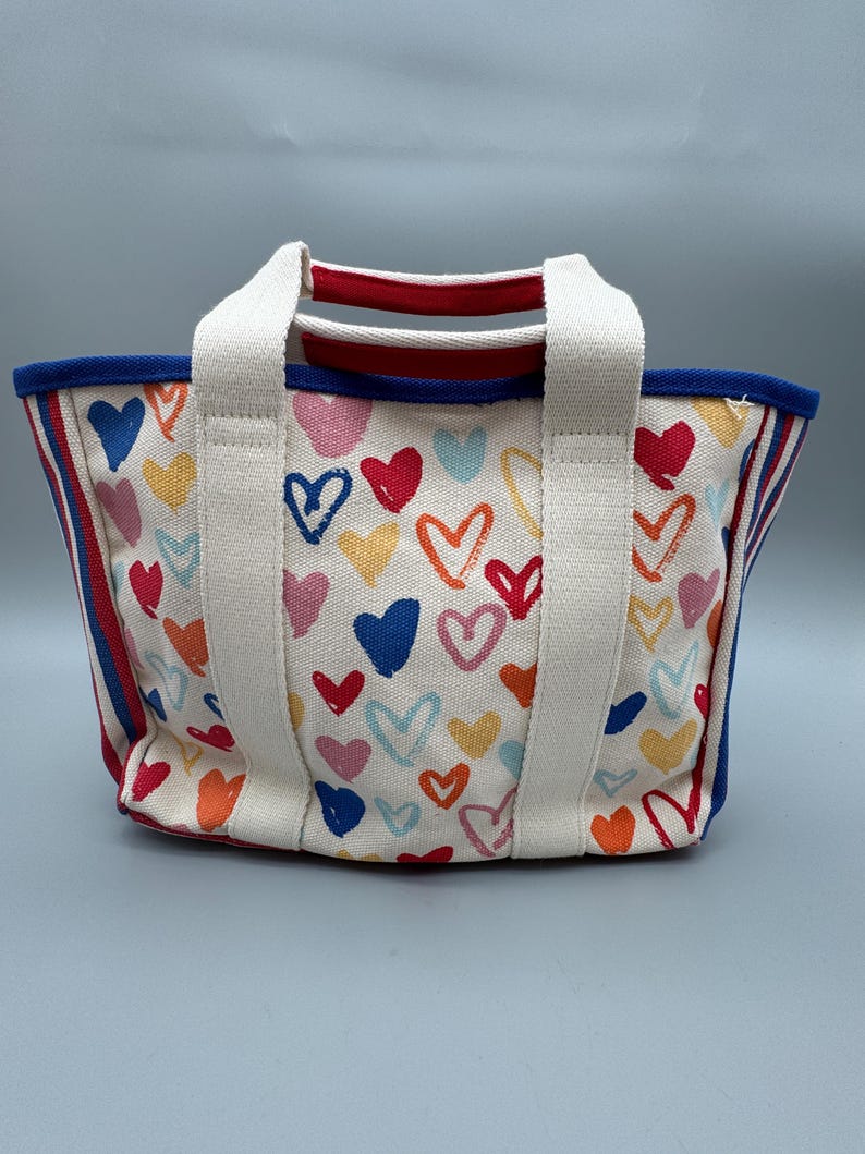 Puede incluir: Un peque&ntilde;o bolso de mano de lona con un estampado de corazones coloridos sobre un fondo blanco. Tiene asas blancas, un ribete azul y rayas rojas y azules en los laterales. Los corazones son de varios colores, como rojo, rosa, azul y amarillo.