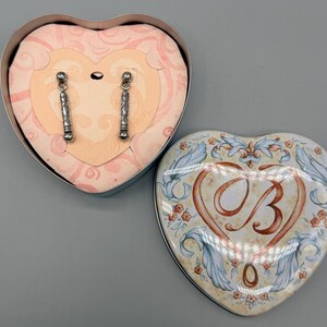 May include: Silver-toned dangle earrings with a geometric design are displayed inside a heart-shaped box. The box is open, revealing the earrings against a pink background. A second heart-shaped box, with a floral design and the letter "B", is next to it.