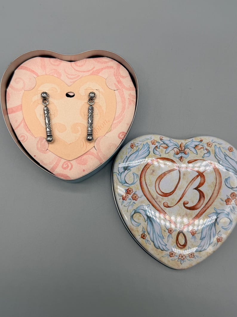 May include: Silver-toned dangle earrings with a geometric design are displayed inside a heart-shaped box. The box is open, revealing the earrings against a pink background. A second heart-shaped box, with a floral design and the letter "B", is next to it.