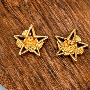 May include: A pair of gold-colored star-shaped stud earrings. Each earring features a five-pointed star design with a detailed center. The earrings are set against a wooden surface.