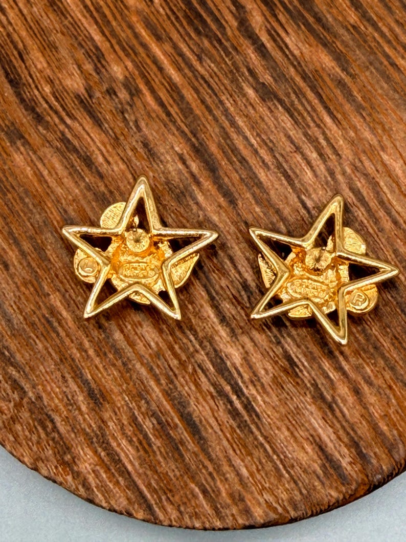 May include: A pair of gold-colored star-shaped stud earrings. Each earring features a five-pointed star design with a detailed center. The earrings are set against a wooden surface.