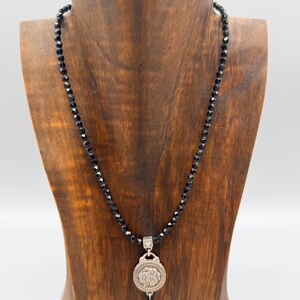 May include: A black beaded necklace with a silver pendant. The pendant features a circular design with a floral pattern. The necklace is displayed on a wooden jewellery stand against a white background.