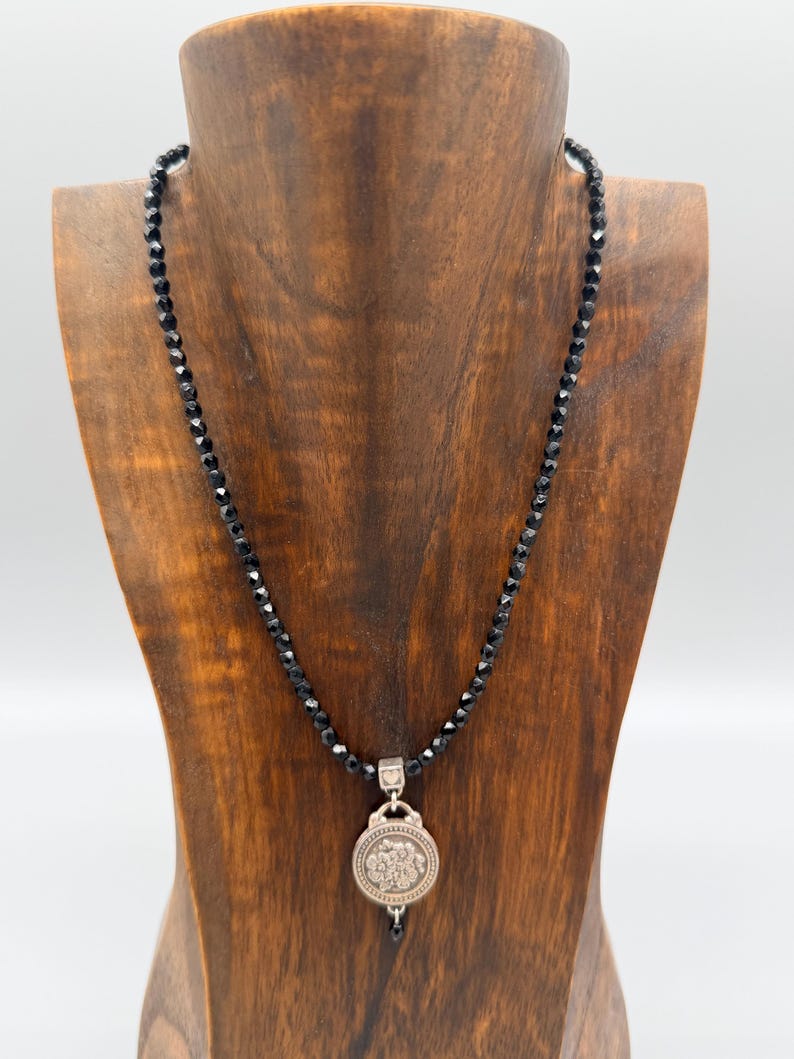 May include: A black beaded necklace with a silver pendant. The pendant features a circular design with a floral pattern. The necklace is displayed on a wooden jewellery stand against a white background.