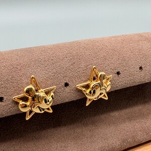 May include: A pair of gold-colored star-shaped stud earrings. Each earring features a Mickey Mouse figure in the center. The earrings are displayed on a brown surface.