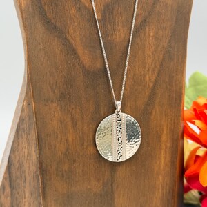 Puede incluir: Un collar plateado con un colgante redondo. El colgante tiene una textura martillada en un lado y un dise&ntilde;o estampado en el otro. El collar se exhibe en un soporte de madera.