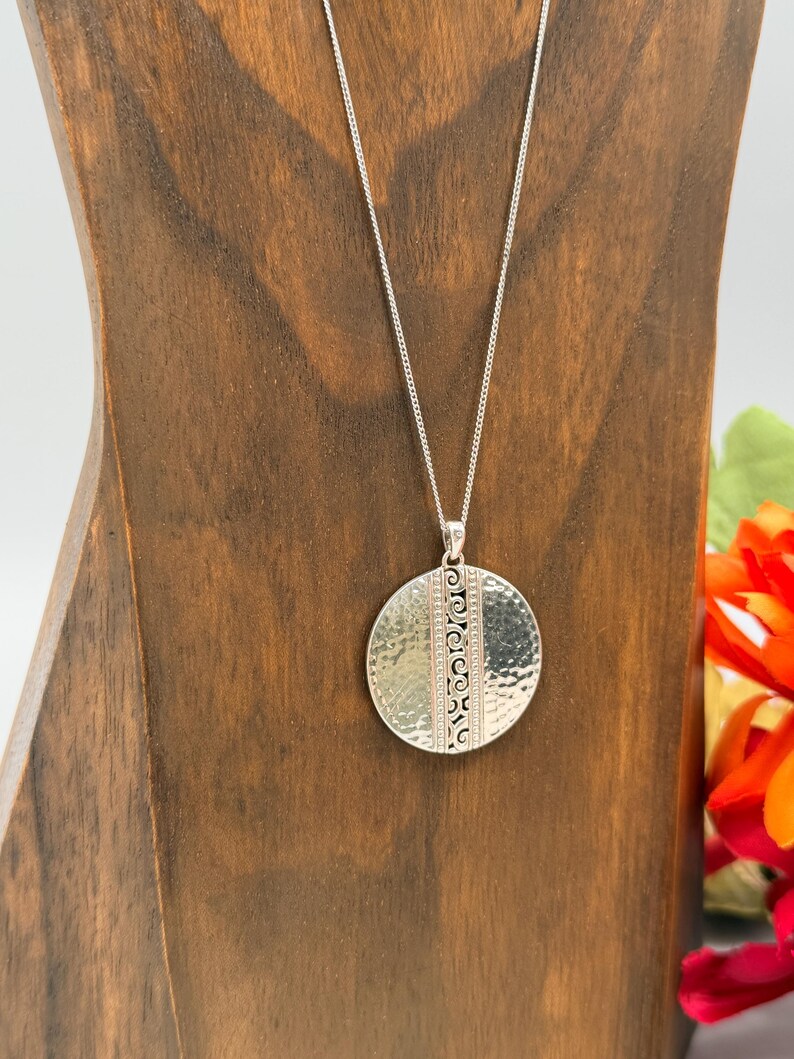 Puede incluir: Un collar plateado con un colgante redondo. El colgante tiene una textura martillada en un lado y un dise&ntilde;o estampado en el otro. El collar se exhibe en un soporte de madera.
