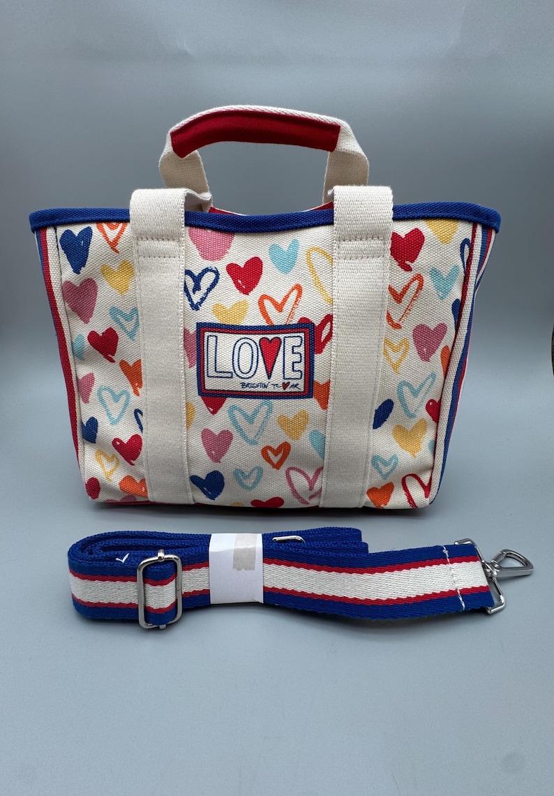 Puede incluir: Un bolso tote de lona con un estampado de corazones coloridos y la palabra "LOVE" en la parte delantera. El bolso tiene un ribete rojo y azul, asas blancas y una correa de hombro a rayas azul y blanca desmontable.