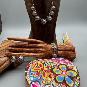 May include: A gold-toned necklace, bracelet, and earrings set with silver and black filigree accents. The jewelry is displayed on a wooden hand and mannequin. A heart-shaped tin with a colorful floral design is in the foreground.