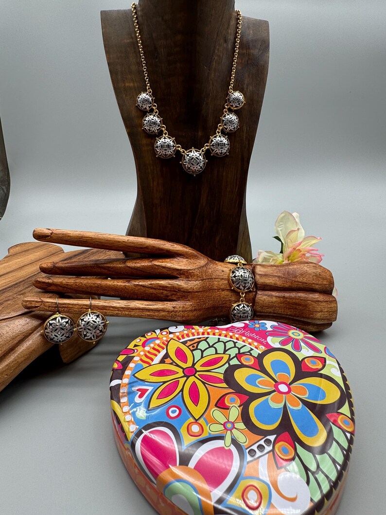 May include: A gold-toned necklace, bracelet, and earrings set with silver and black filigree accents. The jewelry is displayed on a wooden hand and mannequin. A heart-shaped tin with a colorful floral design is in the foreground.