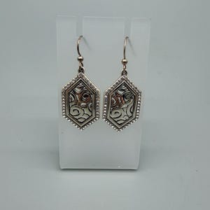 Puede incluir: Un par de pendientes plateados. Cada pendiente tiene forma hexagonal con un dise&ntilde;o calado decorativo en el centro. Los bordes est&aacute;n bordeados con peque&ntilde;os cristales transparentes. Los pendientes cuelgan de un gancho simple.