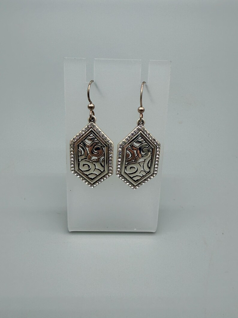 Puede incluir: Un par de pendientes plateados. Cada pendiente tiene forma hexagonal con un dise&ntilde;o calado decorativo en el centro. Los bordes est&aacute;n bordeados con peque&ntilde;os cristales transparentes. Los pendientes cuelgan de un gancho simple.