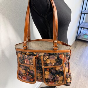 May include: A floral print tote bag with brown leather handles and accents. The bag features a black background with a pattern of roses in shades of orange, yellow, and purple. Two front pockets add to the bag's design.