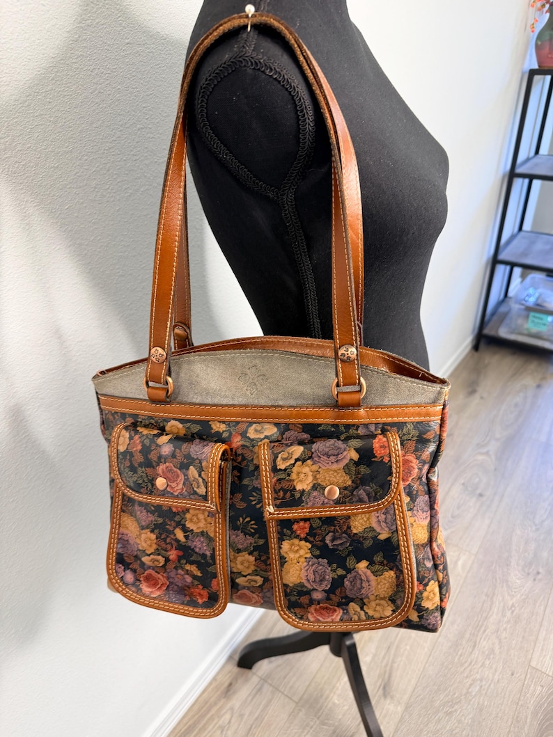 May include: A floral print tote bag with brown leather handles and accents. The bag features a black background with a pattern of roses in shades of orange, yellow, and purple. Two front pockets add to the bag's design.