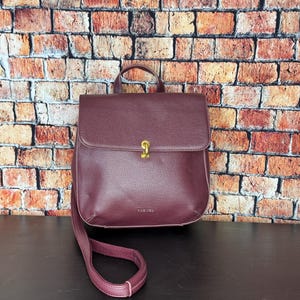 Könnte beinhalten: Ein burgunderfarbener Lederrucksack mit Klappenverschluss und Tragegriff. Die Handtasche hat einen goldfarbenen Verschluss und einen langen, verstellbaren Schulterriemen. Der Hintergrund ist eine Backsteinmauer.