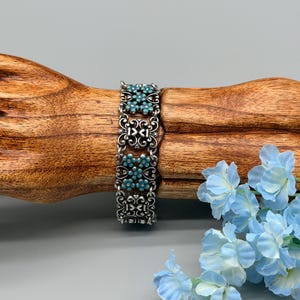 以下が含まれることがあります： ターコイズ色の花のアクセントが付いたシルバーカラーのブレスレット。ブレスレットは、花のパターンの中に小さな丸いターコイズ色の石がセットされた、装飾的でオープンワークのデザインが特徴です。ブレスレットは木製の表面に表示されています。
