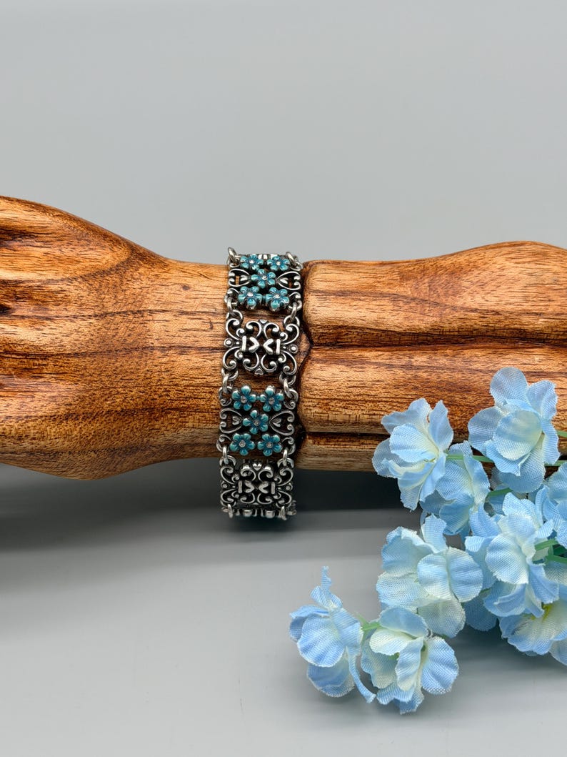 以下が含まれることがあります： ターコイズ色の花のアクセントが付いたシルバーカラーのブレスレット。ブレスレットは、花のパターンの中に小さな丸いターコイズ色の石がセットされた、装飾的でオープンワークのデザインが特徴です。ブレスレットは木製の表面に表示されています。