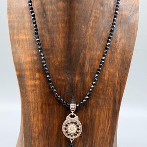 May include: A black beaded necklace with a decorative pendant. The pendant has a silver-tone frame and a central design of small, sparkling stones. The necklace is displayed on a wooden bust.