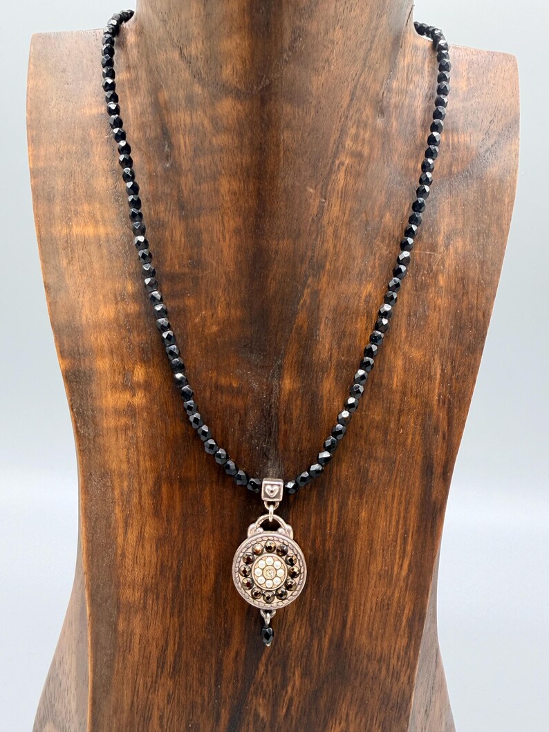 May include: A black beaded necklace with a decorative pendant. The pendant has a silver-tone frame and a central design of small, sparkling stones. The necklace is displayed on a wooden bust.