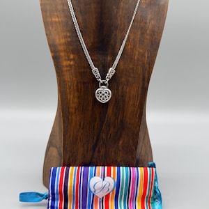 Puede incluir: Un collar plateado con un colgante en forma de coraz&oacute;n se exhibe sobre un busto de madera. El collar tiene una cadena tejida. Una bolsa a rayas colorida con un bot&oacute;n en forma de coraz&oacute;n est&aacute; en la base del busto.