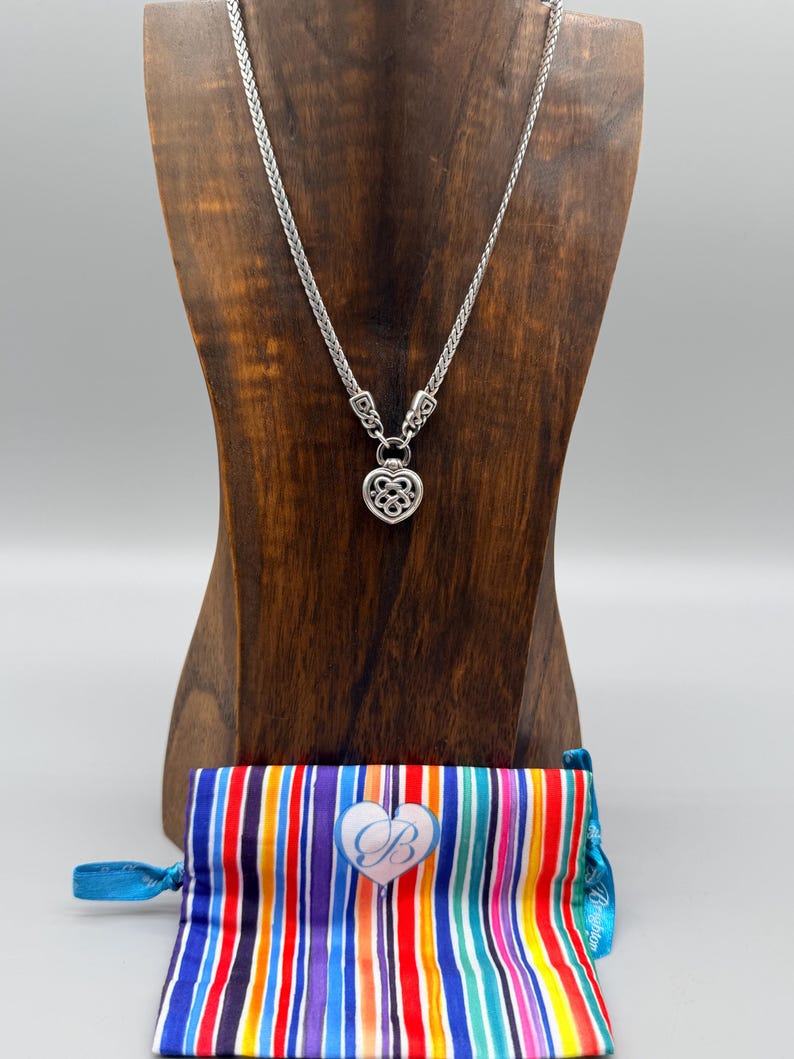 Puede incluir: Un collar plateado con un colgante en forma de coraz&oacute;n se exhibe sobre un busto de madera. El collar tiene una cadena tejida. Una bolsa a rayas colorida con un bot&oacute;n en forma de coraz&oacute;n est&aacute; en la base del busto.