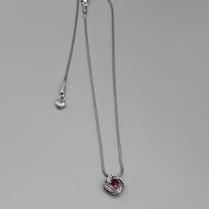 May include: A silver necklace featuring a pendant with a central red gemstone surrounded by a halo of smaller, clear stones. The pendant is square-shaped with rounded edges. The chain is delicate and silver, with a small heart-shaped charm near the clasp.
