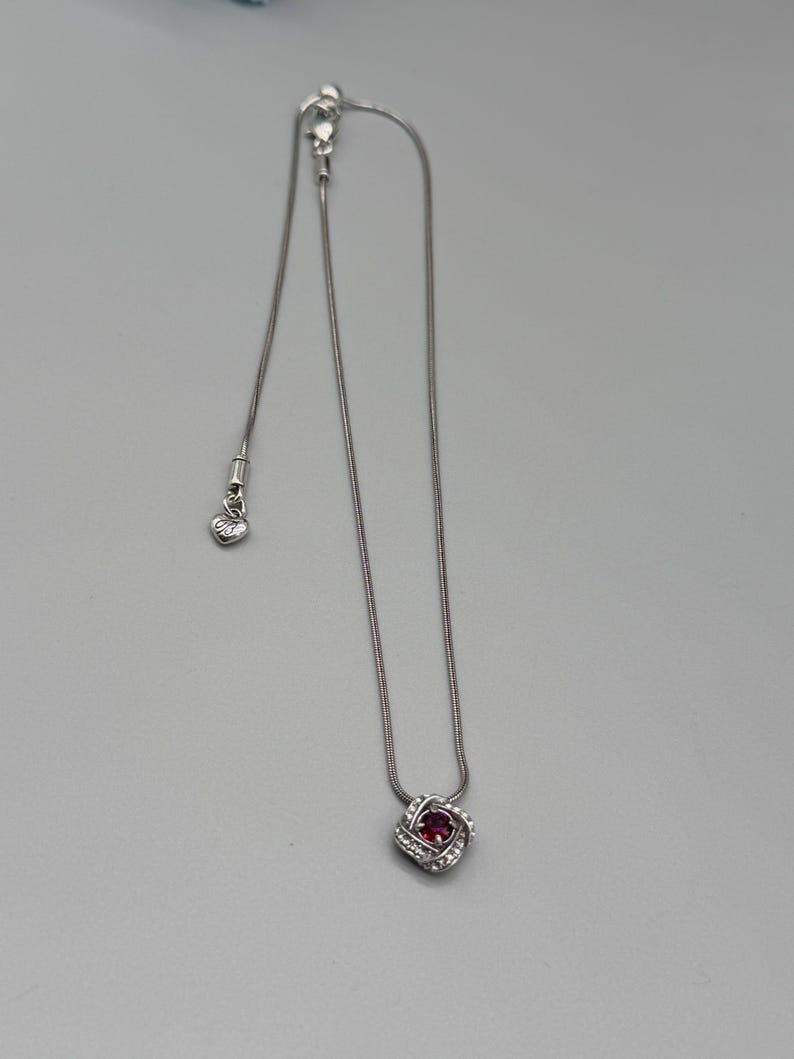 May include: A silver necklace featuring a pendant with a central red gemstone surrounded by a halo of smaller, clear stones. The pendant is square-shaped with rounded edges. The chain is delicate and silver, with a small heart-shaped charm near the clasp.