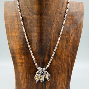 May include: A silver chain necklace featuring three small, dangling charms. Each charm has a textured silver border surrounding a central, smooth, gold-toned inlay. The charms are shaped like an oval, a rectangle, and a triangle, hanging from a braided chain.