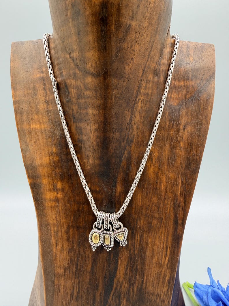 May include: A silver chain necklace featuring three small, dangling charms. Each charm has a textured silver border surrounding a central, smooth, gold-toned inlay. The charms are shaped like an oval, a rectangle, and a triangle, hanging from a braided chain.