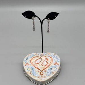 May include: Silver-toned dangle earrings with a geometric design are displayed on a black stand. The earrings are presented on a heart-shaped tin with a floral and scroll design, and a large letter "B" in the center.
