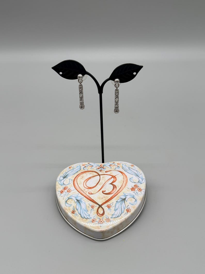 May include: Silver-toned dangle earrings with a geometric design are displayed on a black stand. The earrings are presented on a heart-shaped tin with a floral and scroll design, and a large letter "B" in the center.