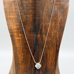 May include: A silver necklace with a small, square pendant featuring a lattice design. The necklace is displayed on a wooden jewelry stand against a white background. The pendant appears to be made of silver or a silver-toned metal.