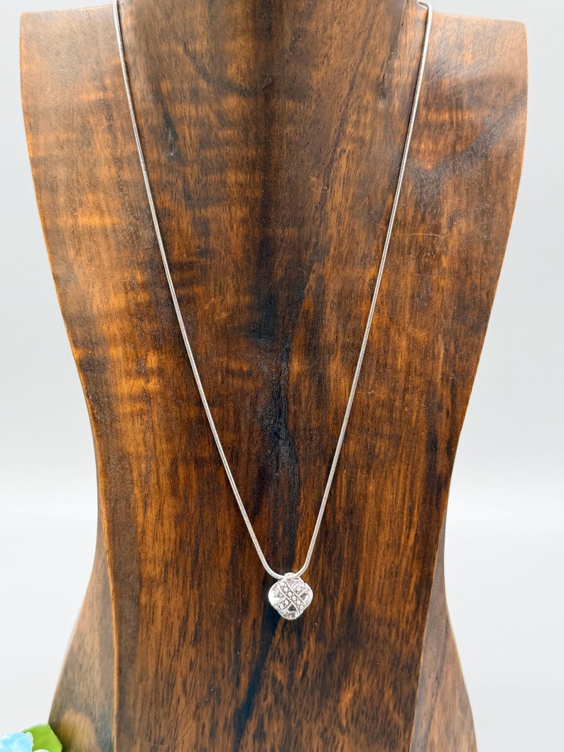 May include: A silver necklace with a small, square pendant featuring a lattice design. The necklace is displayed on a wooden jewelry stand against a white background. The pendant appears to be made of silver or a silver-toned metal.