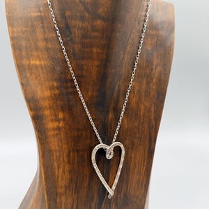 Pu&ograve; includere: Una collana argentata con un ciondolo a forma di cuore ornato da piccole pietre preziose trasparenti. La collana &egrave; esposta su un busto di legno. Il design del ciondolo &egrave; evidenziato dalla delicata catena. Un elegante gioiello.
