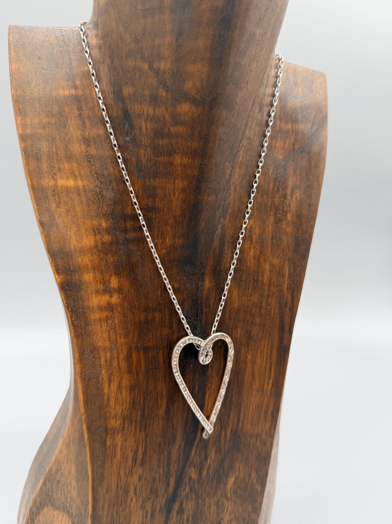 Pu&ograve; includere: Una collana argentata con un ciondolo a forma di cuore ornato da piccole pietre preziose trasparenti. La collana &egrave; esposta su un busto di legno. Il design del ciondolo &egrave; evidenziato dalla delicata catena. Un elegante gioiello.