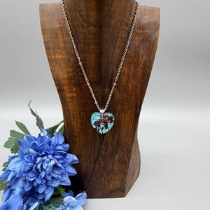 May include: A silver-toned necklace with a heart-shaped pendant. The pendant features a floral design with the word "love" and is displayed on a wooden jewelry stand. Blue flowers are in the foreground.